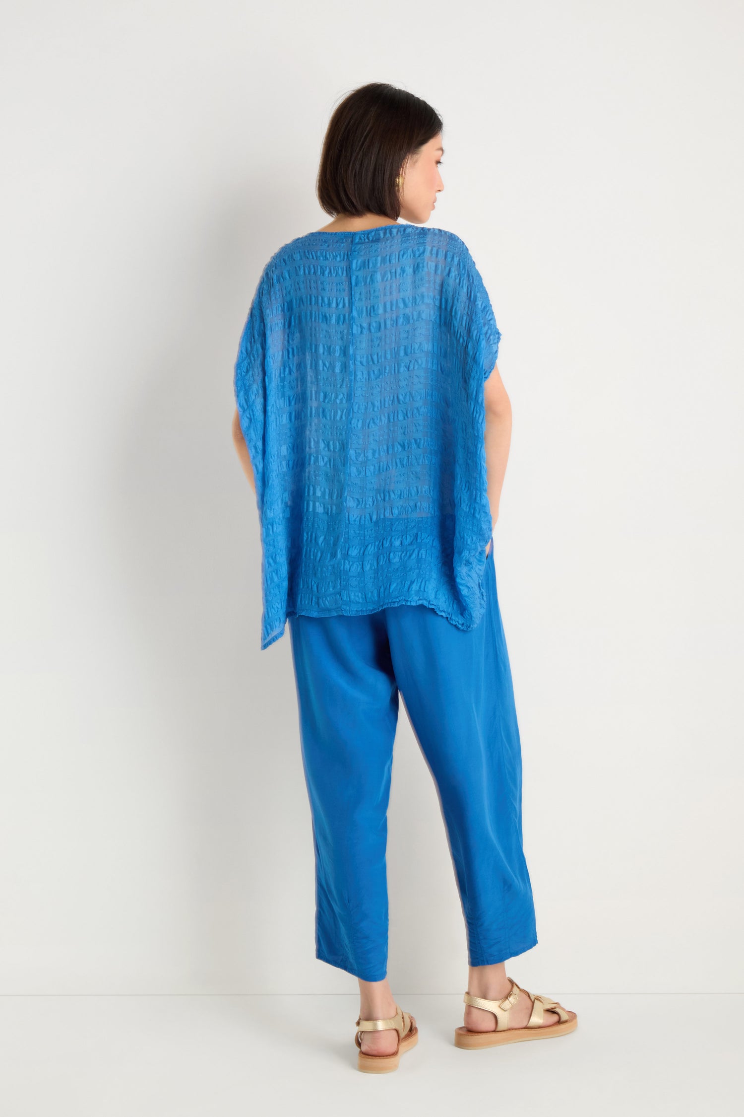 A woman stands facing away, modeling the Sheer Crinkle Silk Boxy Top in blue with matching pants and beige sandals, set against a plain white background.