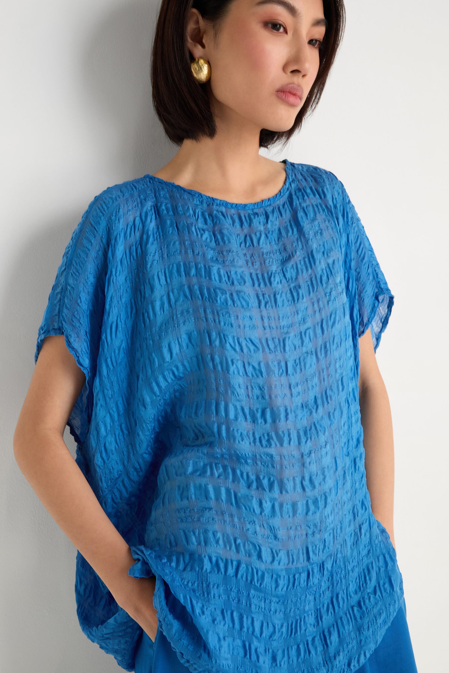 A woman wears the Sheer Crinkle Silk Boxy Top, a lightweight, loose-fitting blue blouse with short sleeves, paired with gold earrings and blue pants, standing against a white background.