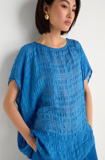 A woman wears the Sheer Crinkle Silk Boxy Top, a lightweight, loose-fitting blue blouse with short sleeves, paired with gold earrings and blue pants, standing against a white background.