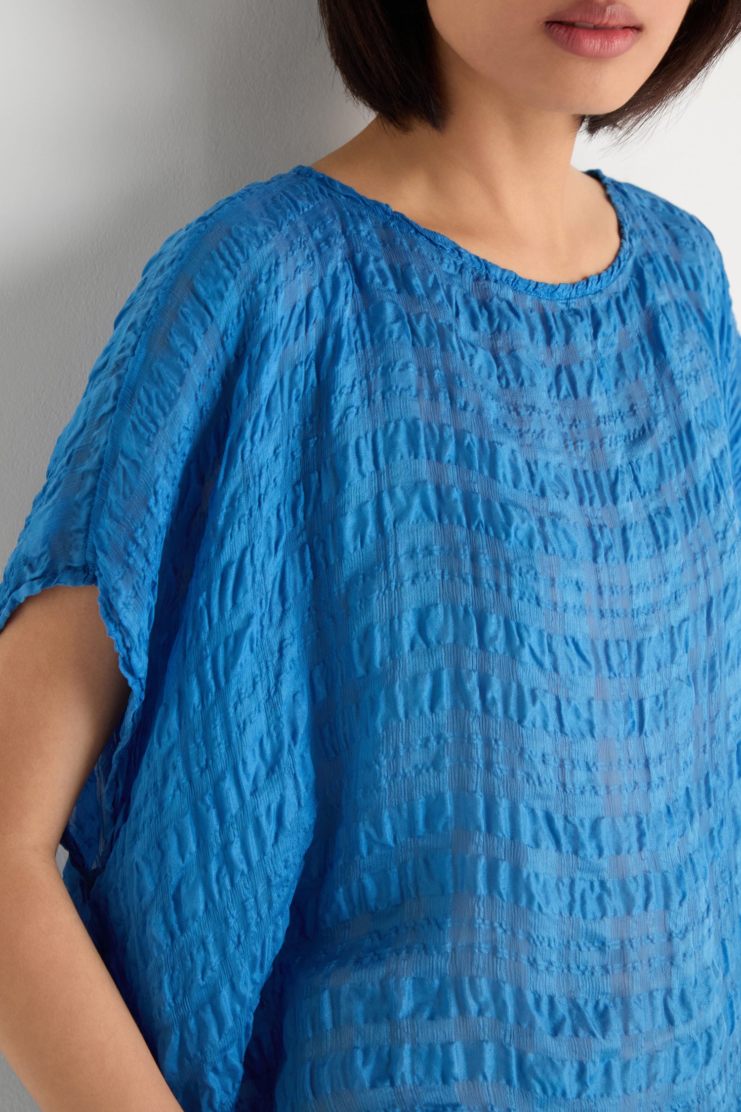 A woman wears the Sheer Crinkle Silk Boxy Top, a lightweight, semi-sheer blue short-sleeve piece, against a light-colored wall.