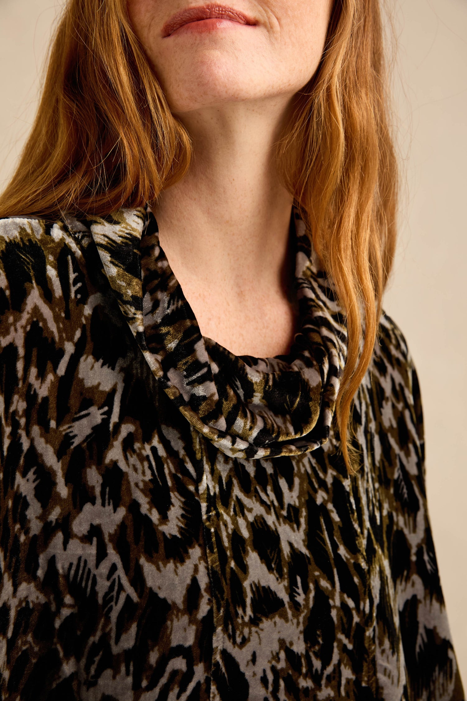 A woman with long auburn hair wears the Animal Print Cowl Neck Velvet Top, crafted from a silk-blend velvet and featuring a striking black, gray, and green abstract pattern.
