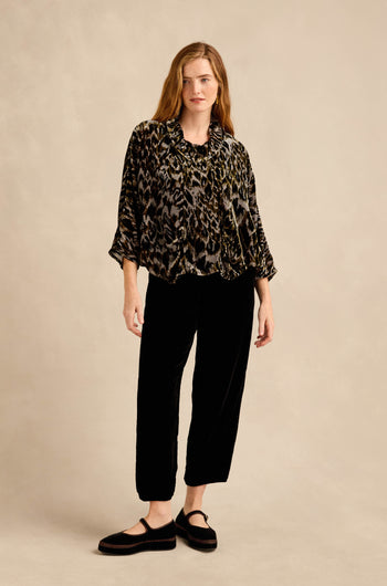 Against a beige background, a woman with long, wavy hair and a neutral expression wears the Animal Print Cowl Neck Velvet Top, styled with black velvet pants and black shoes.
