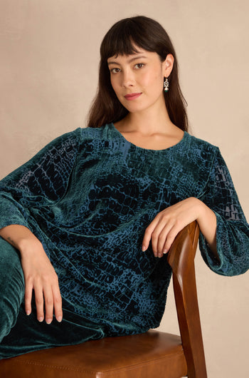 Woman with dark hair sits on a brown chair, wearing the Deep Turquoise Leopard Devore Layered top, looking at the camera with a neutral expression.