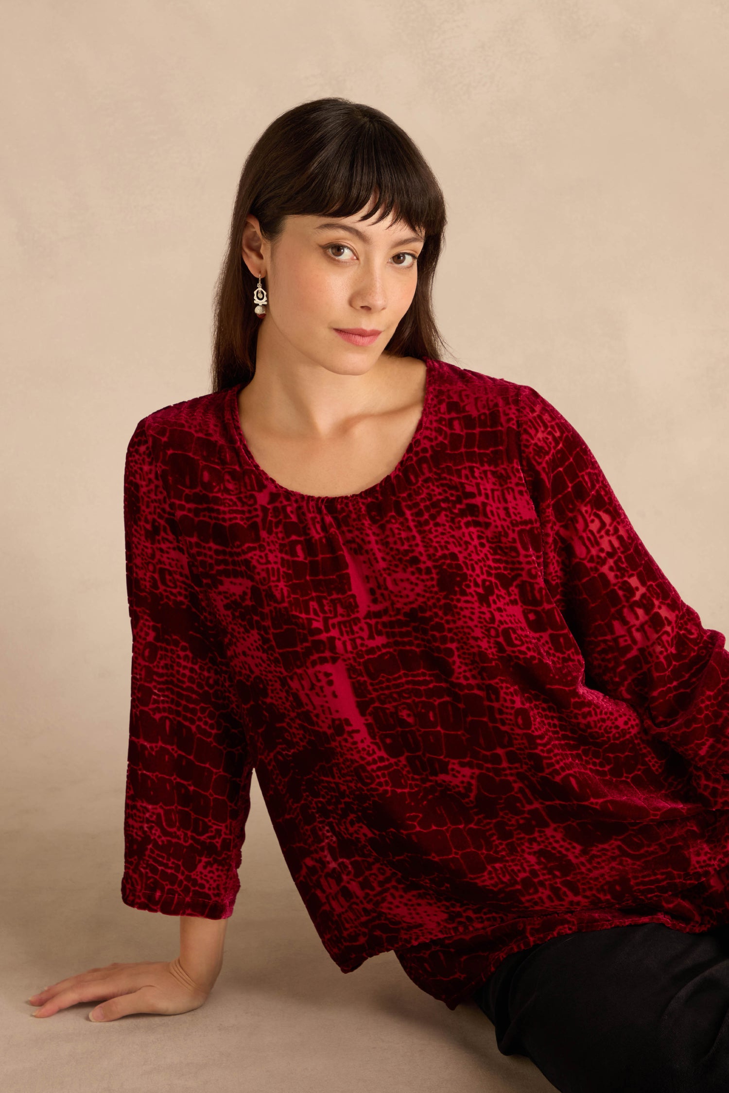 Woman with straight brown hair and bangs, wearing the Ruby Leopard Devore Layered Top and earrings, sits against a beige background, looking at the camera neutrally.