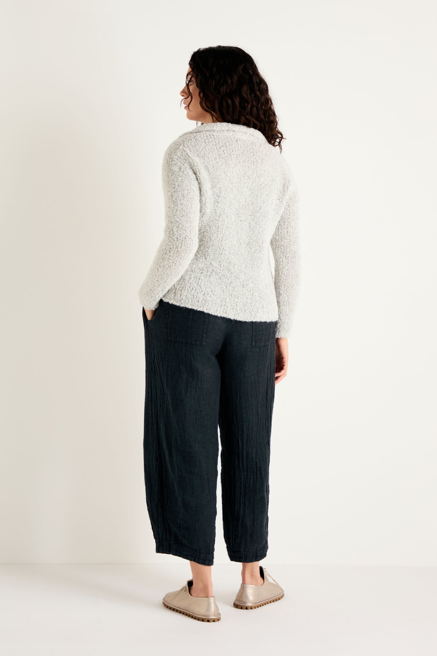 A person stands with their back to the camera, wearing the Boucle Collar Cardigan—a warm knit sweater with a subtle boucle texture—paired with loose black pants and beige slip-on shoes against a plain white background.