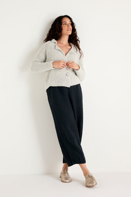 A woman stands against a white wall, wearing the Boucle Collar Cardigan—a light gray wool alpaca cardigan with a subtle boucle texture—paired with black wide-leg pants and beige shoes. Her hands are in her pockets as she looks to the side.