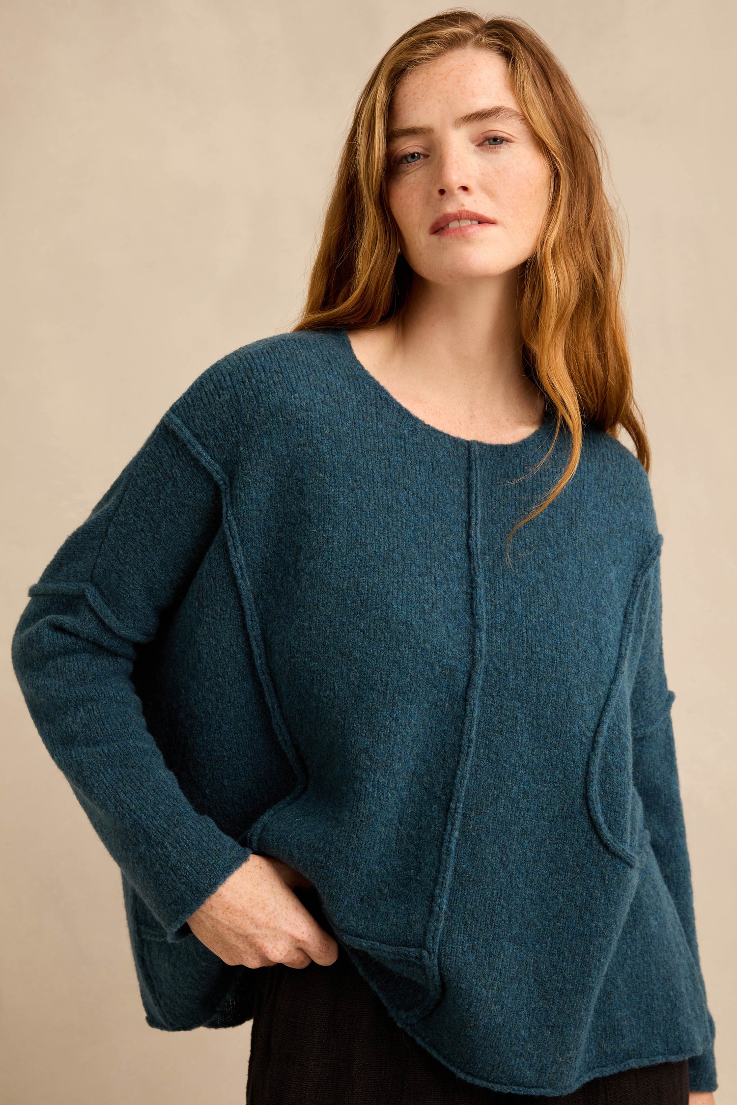 A woman with long red hair wears the Deep Turquoise Seamed Knit—a loose-fitting, teal wool-rich jumper with curved seam detailing and practical pockets—as she stands against a neutral background.