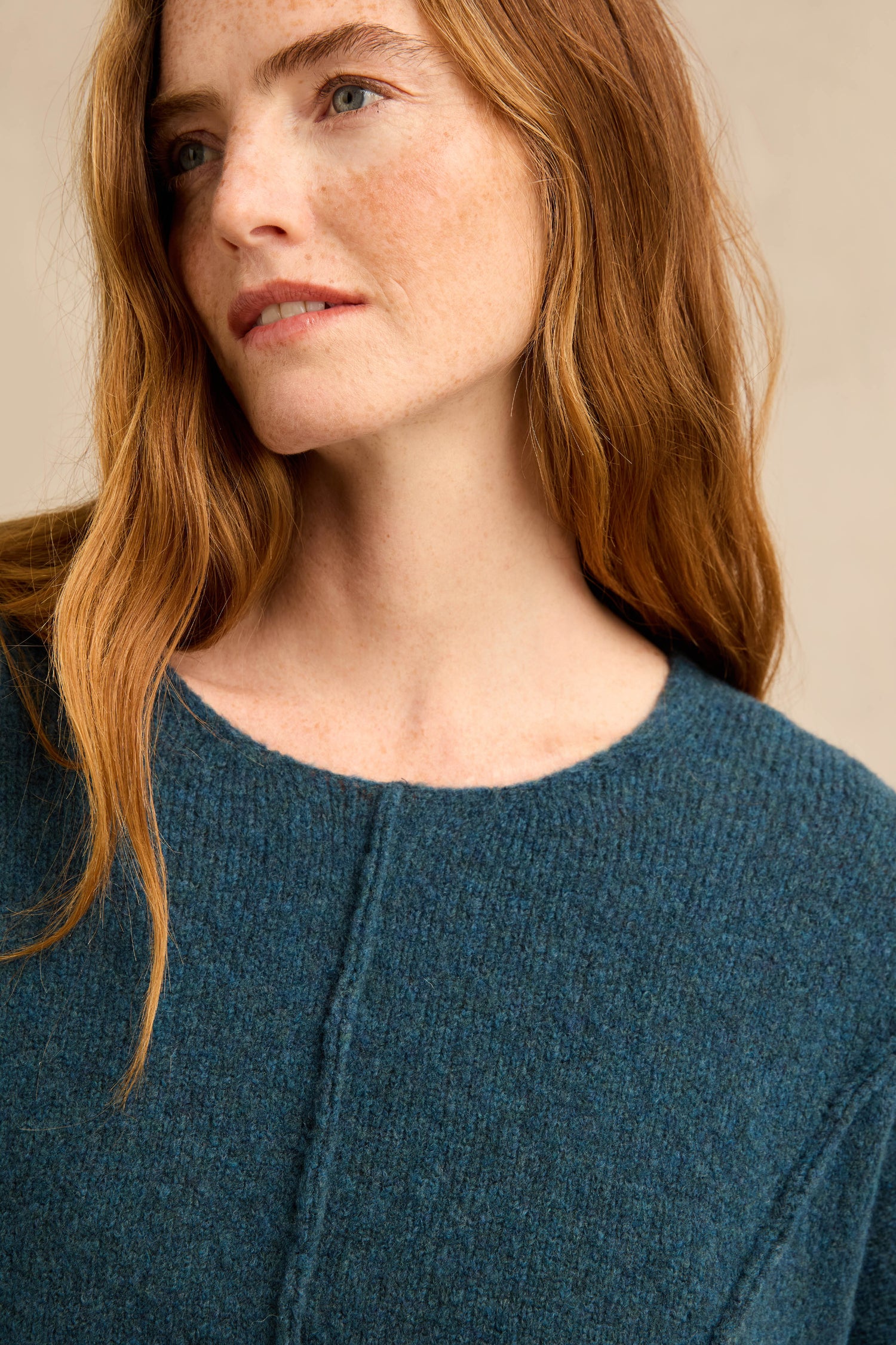 A woman with long red hair wears the Deep Turquoise Seamed Knit—a textured blue wool-rich jumper with subtle curved seam detailing—and looks slightly to the side against a neutral background.