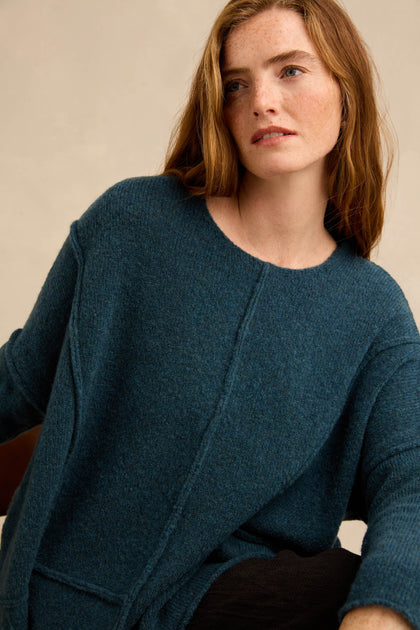 A woman with long auburn hair wears the Deep Turquoise Seamed Knit—a textured blue sweater featuring curved seam detailing—as she sits, looking slightly to the side against a neutral background.