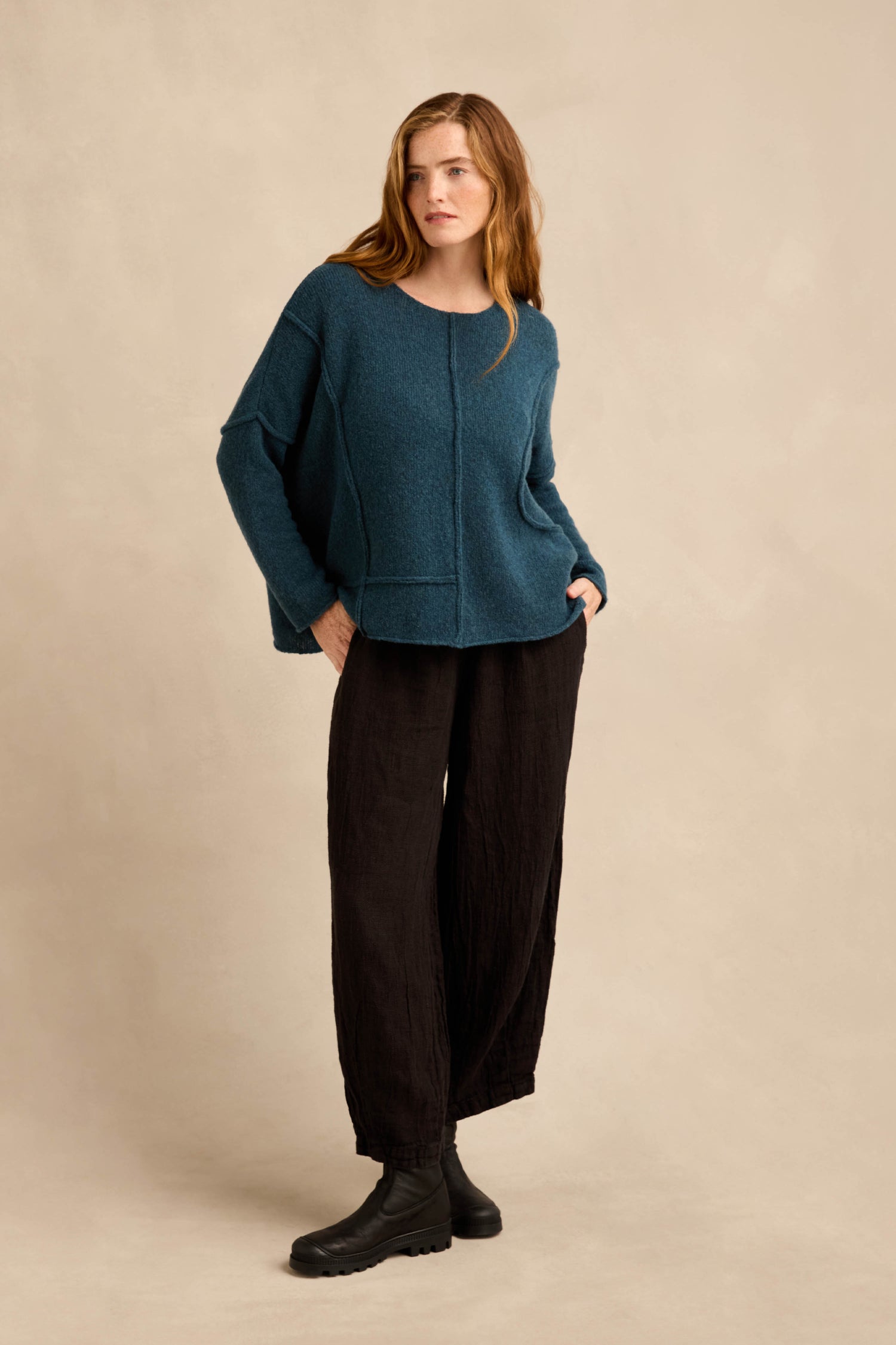 A person stands against a beige background wearing the Deep Turquoise Seamed Knit with curved seam detailing, paired with loose black pants and black boots, hands in pockets and looking slightly to the side.