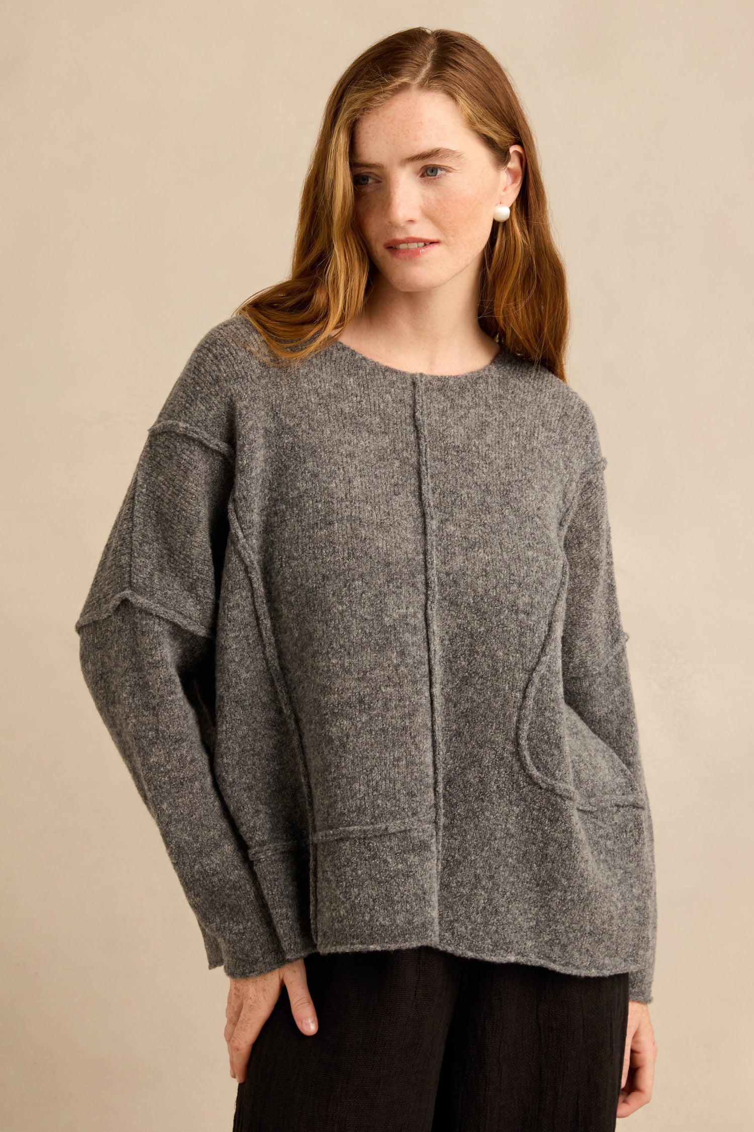 A woman is wearing the Grey Seamed Knit—a loose-fitting, long-sleeved gray wool-rich jumper with curved seam detailing—paired with black pants, standing against a beige background.