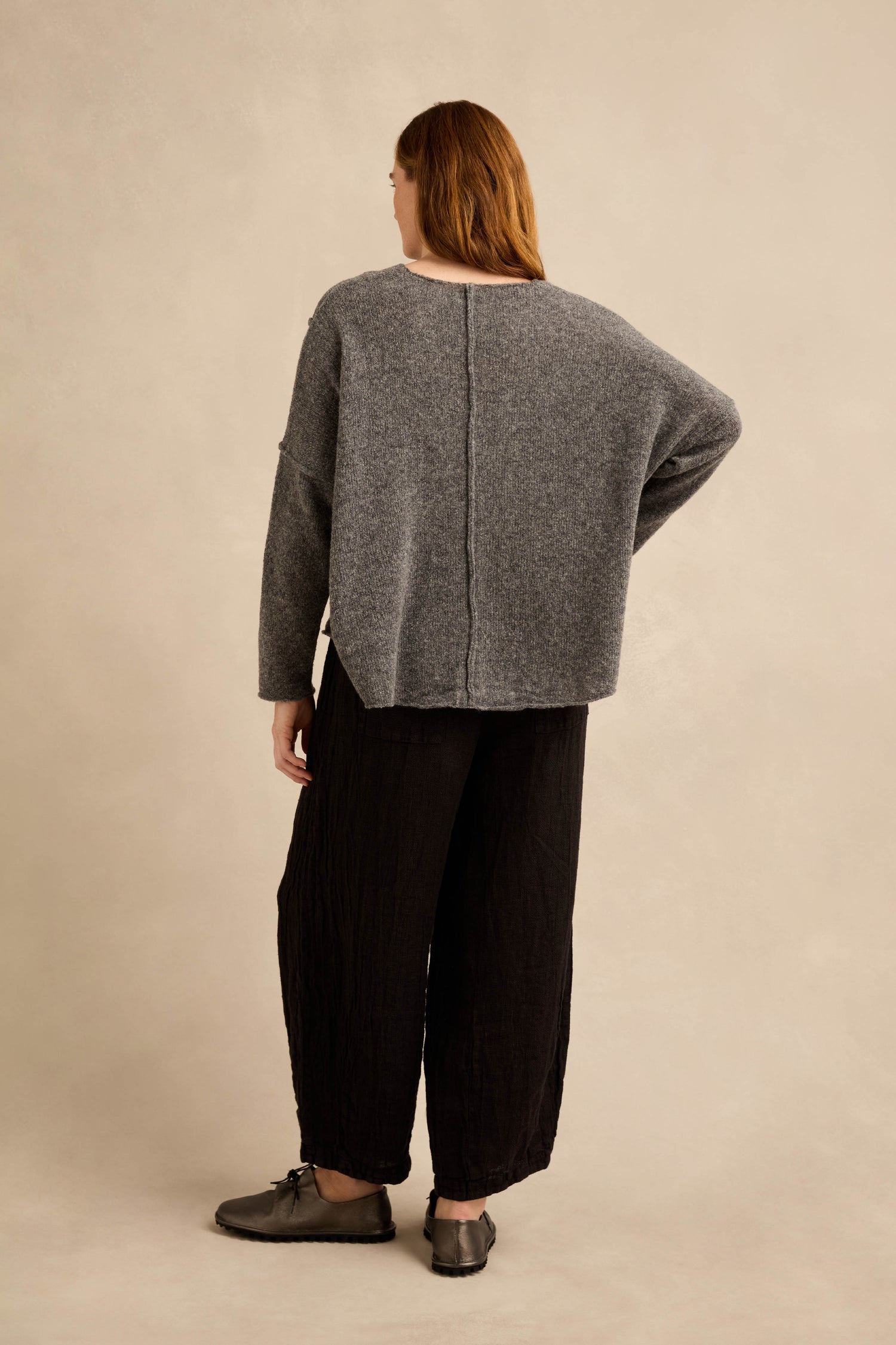 Person standing with back to camera, wearing the Grey Seamed Knit—a loose gray wool-rich jumper—paired with black wide-leg pants and gray shoes, against a plain beige background.