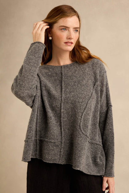 A woman stands against a neutral background, looking to the side with one hand touching her hair, wearing the Grey Seamed Knit sweater paired with black pants.