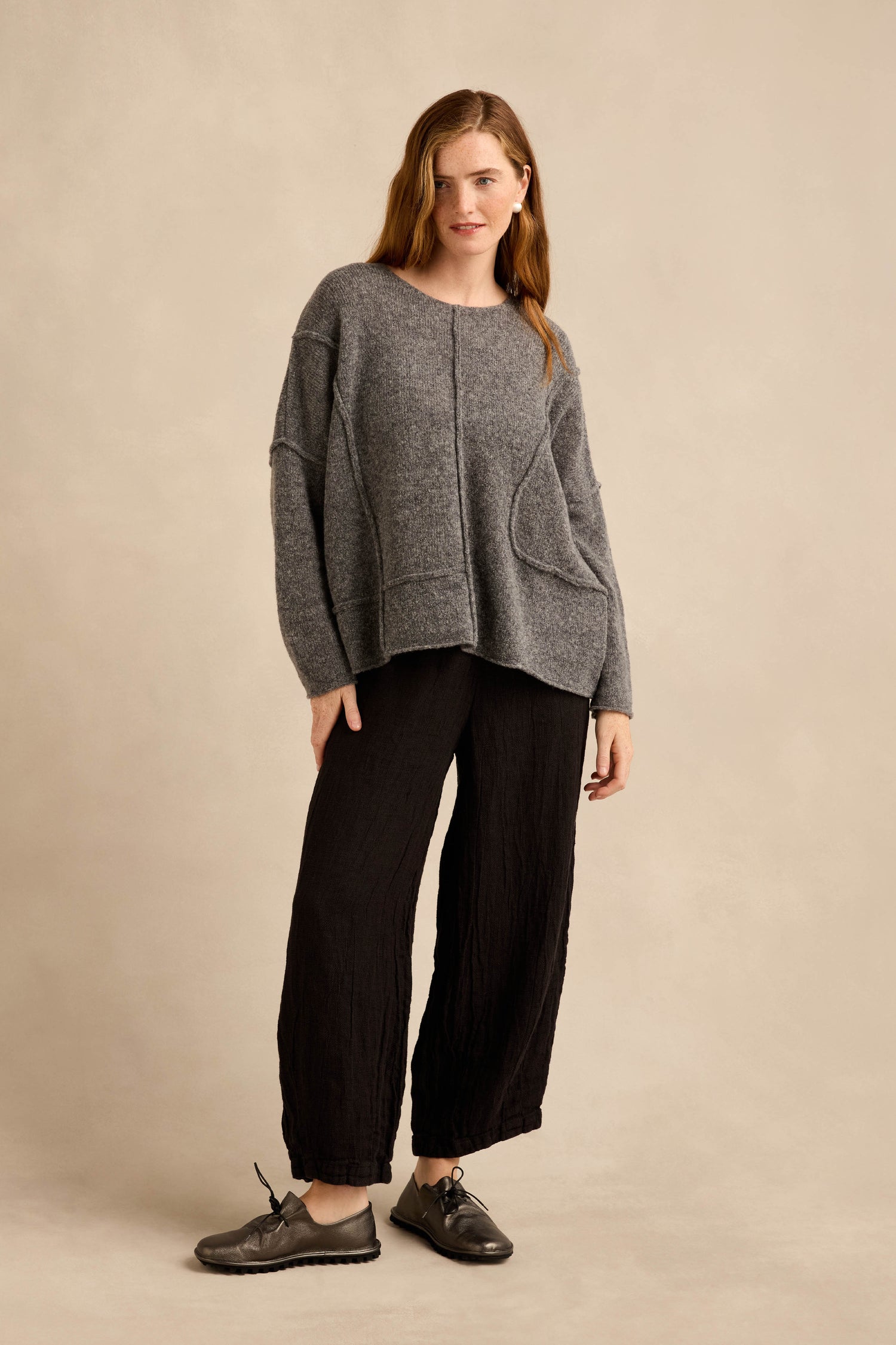 A woman stands against a beige background wearing the Grey Seamed Knit sweater—a relaxed wool knit in grey with curved seam detailing—paired with black wide-leg pants and black shoes.