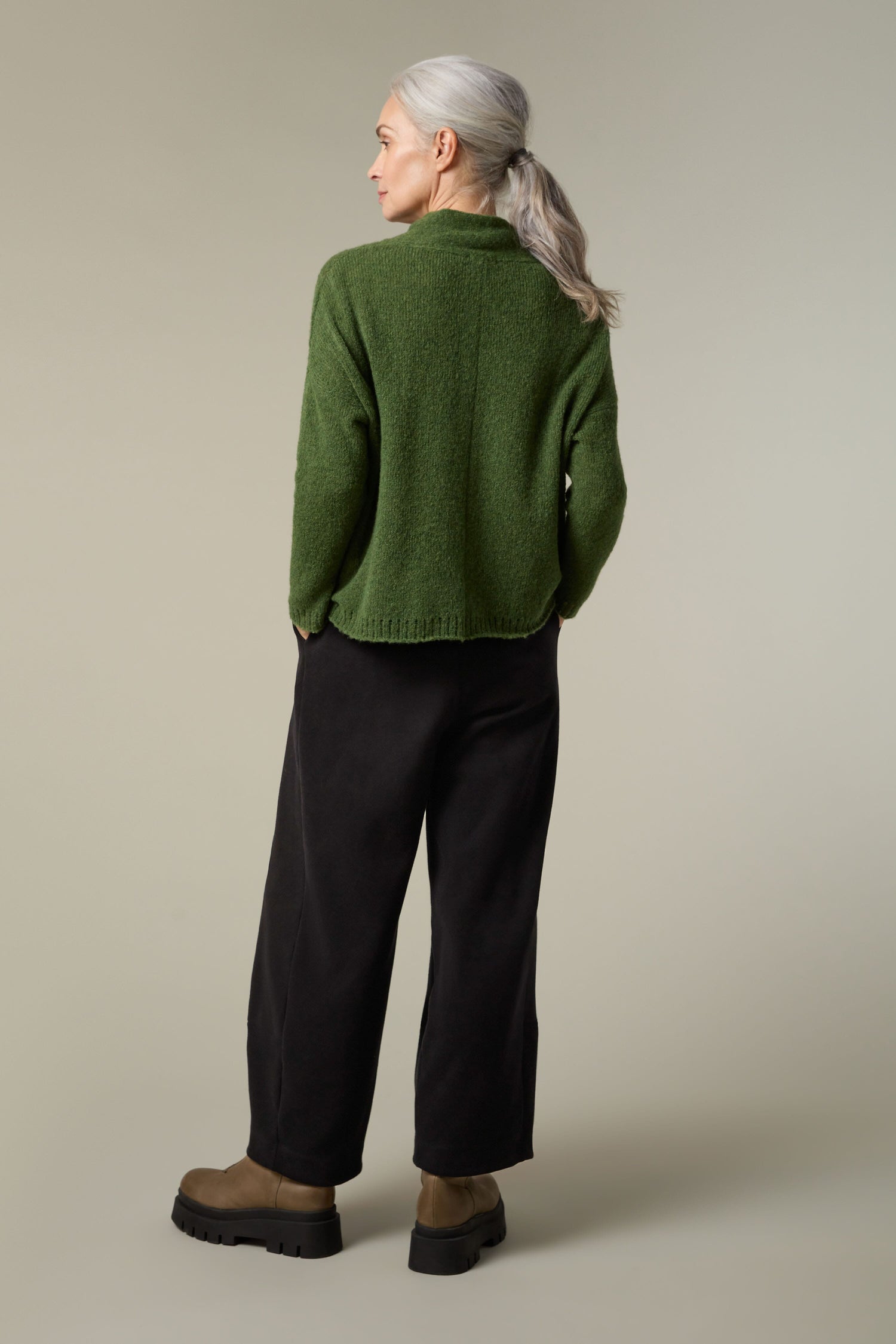 The back view of a woman wearing a cosy green Panel Knit sweater and black pants.