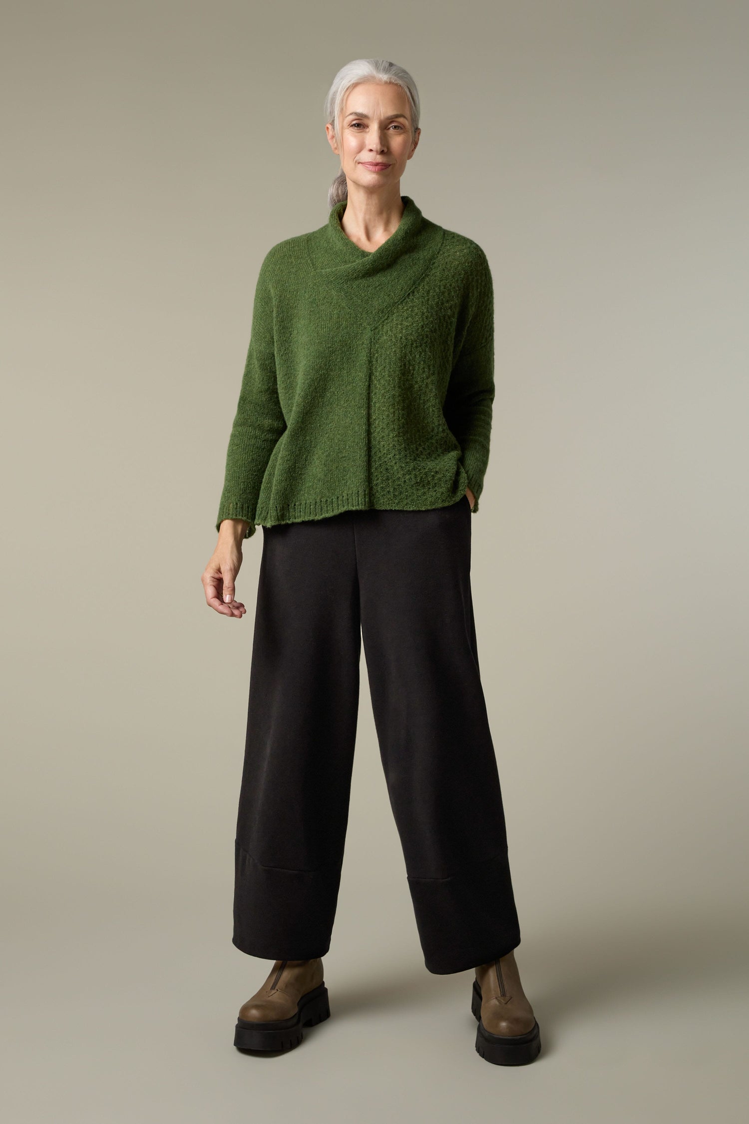 A woman wearing a Grizas Panel Knit green sweater, creating a cosy layering look with her black wide leg pants.