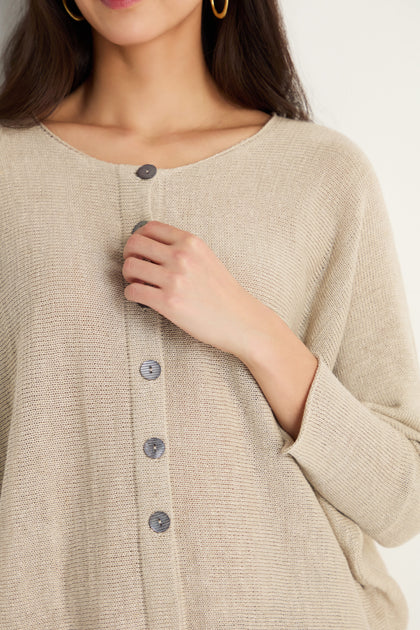 A woman, face partially visible, wears the Boxy Linen Button Cardigan in beige with gray buttons, holding one near her chest. This lightweight layering piece adds effortless style to any look.