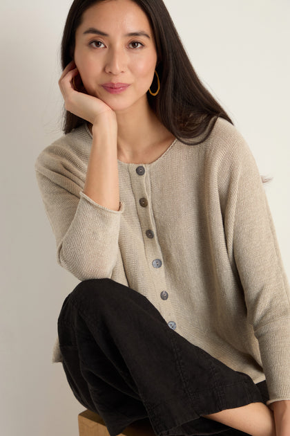 A woman with long dark hair sits on a stool, wearing the Boxy Linen Button Cardigan and black pants. With one hand resting on her cheek, she shows off this lightweight layering piece—ideal for any season.