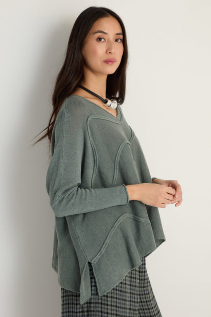 Woman with long dark hair and a neutral expression stands against a white wall, wearing the Swirl Seamed Linen Knit Top in gray, paired with a plaid skirt and chunky necklace for a relaxed silhouette.