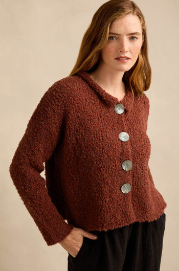 A woman stands against a plain beige background wearing the Three Button Cardigan, an oversized textured dark red knit featuring large silver buttons, paired with black pants.