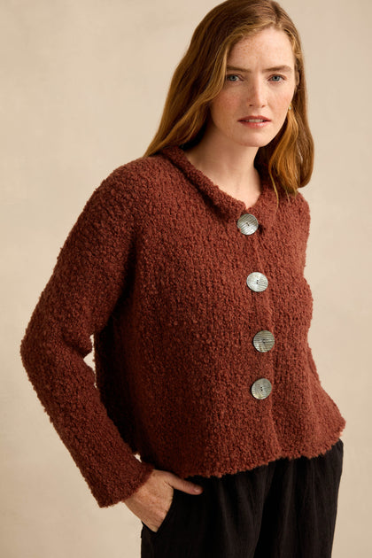 A woman stands against a plain beige background wearing the Three Button Cardigan, an oversized textured dark red knit featuring large silver buttons, paired with black pants.