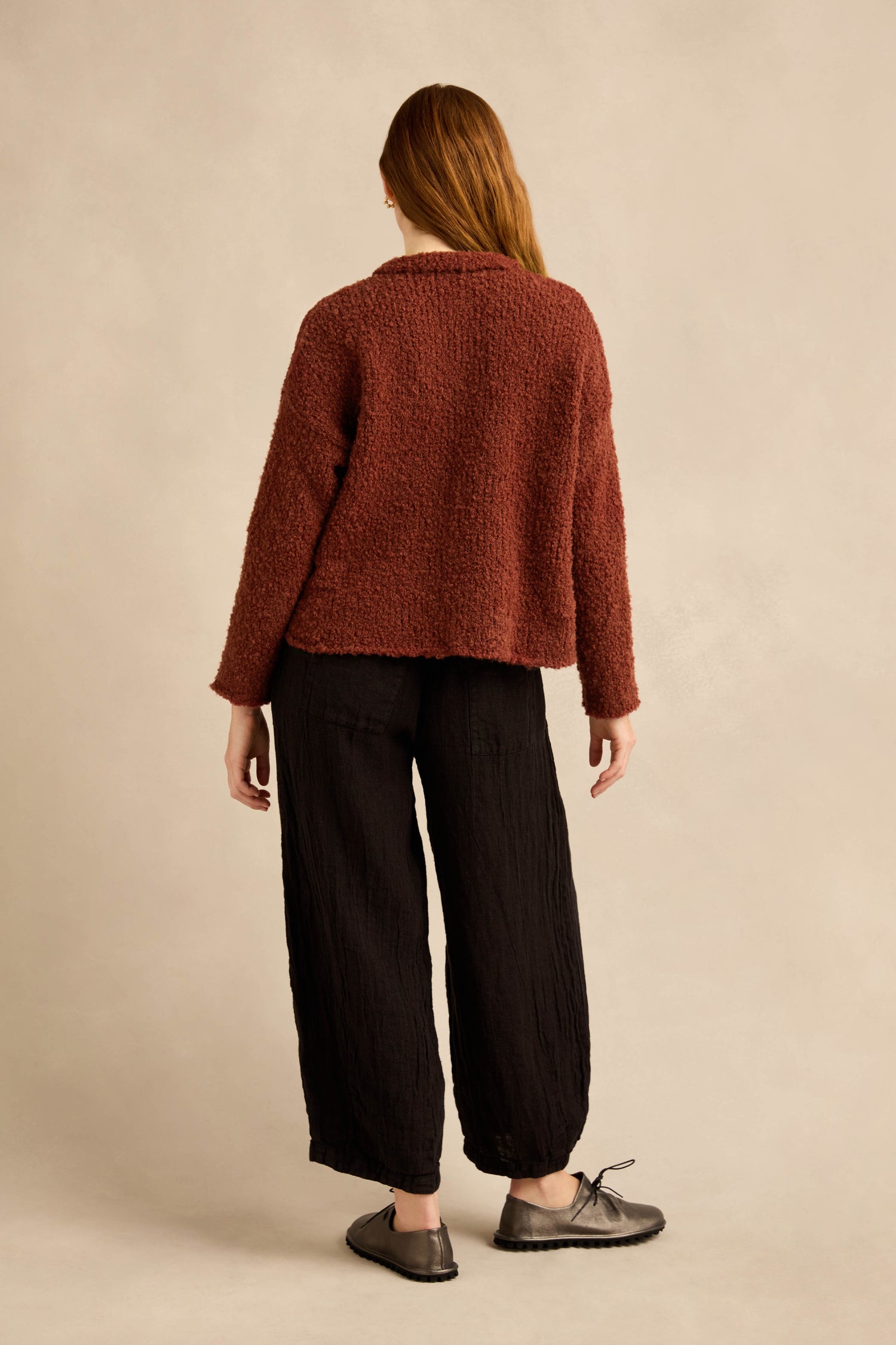 A person with long hair, wearing the Three Button Cardigan in maroon with a boxy knitwear fit, loose black pants, and gray shoes, stands facing away from the camera against a neutral background.