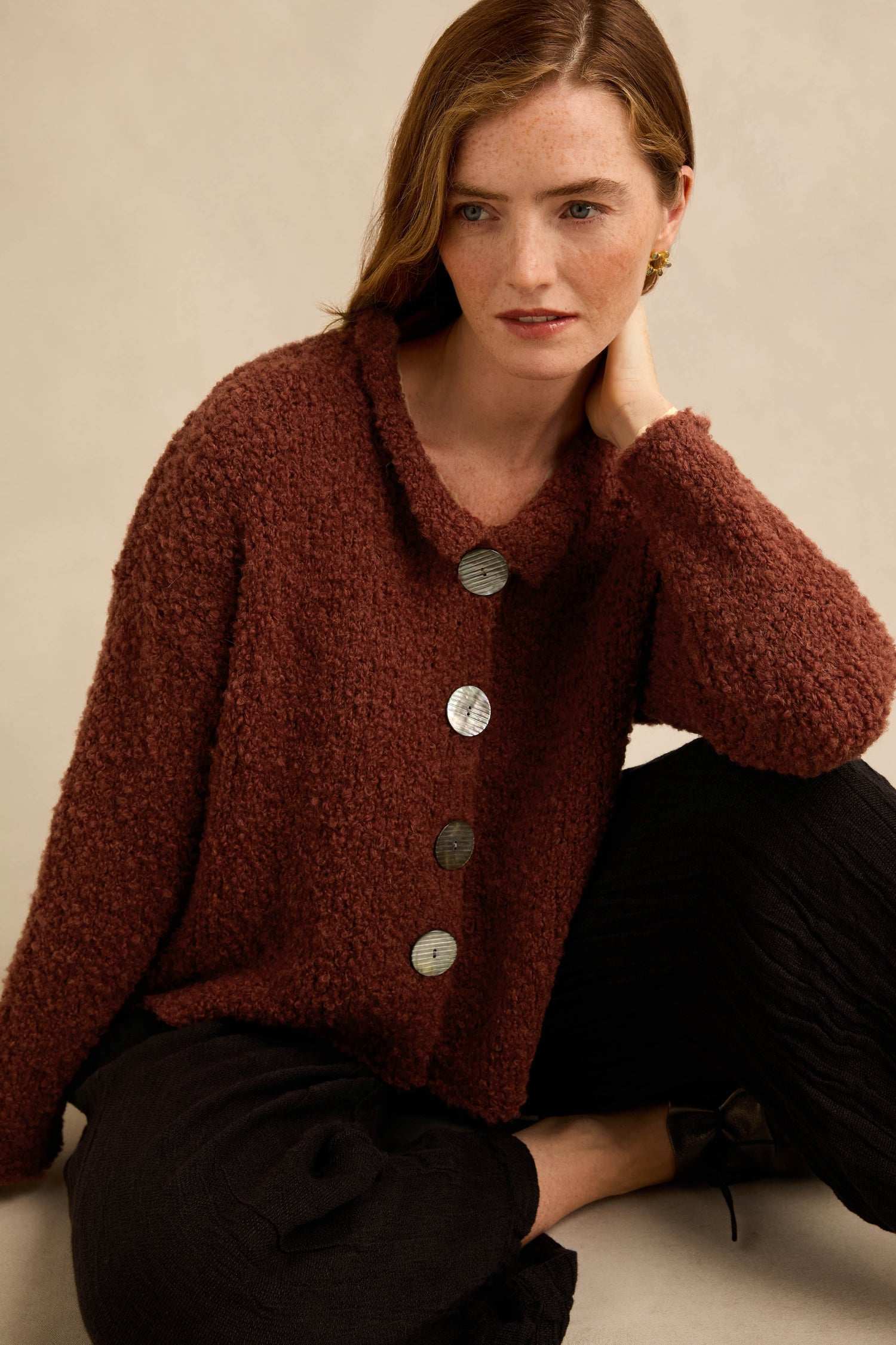 A woman with light skin and brown hair wears the Three Button Cardigan—a textured maroon Grizas knitwear piece featuring large buttons—paired with black pants, sitting with one hand resting on her neck against a neutral background.