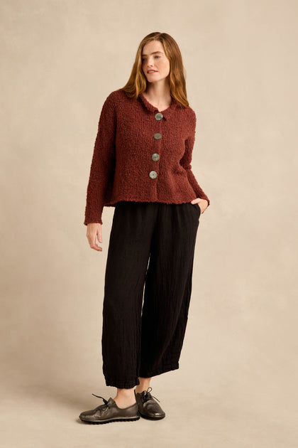 A woman stands against a beige background wearing the Three Button Cardigan, an oversized piece in textured burgundy knit, paired with loose black pants and black shoes, with one hand in her pocket.