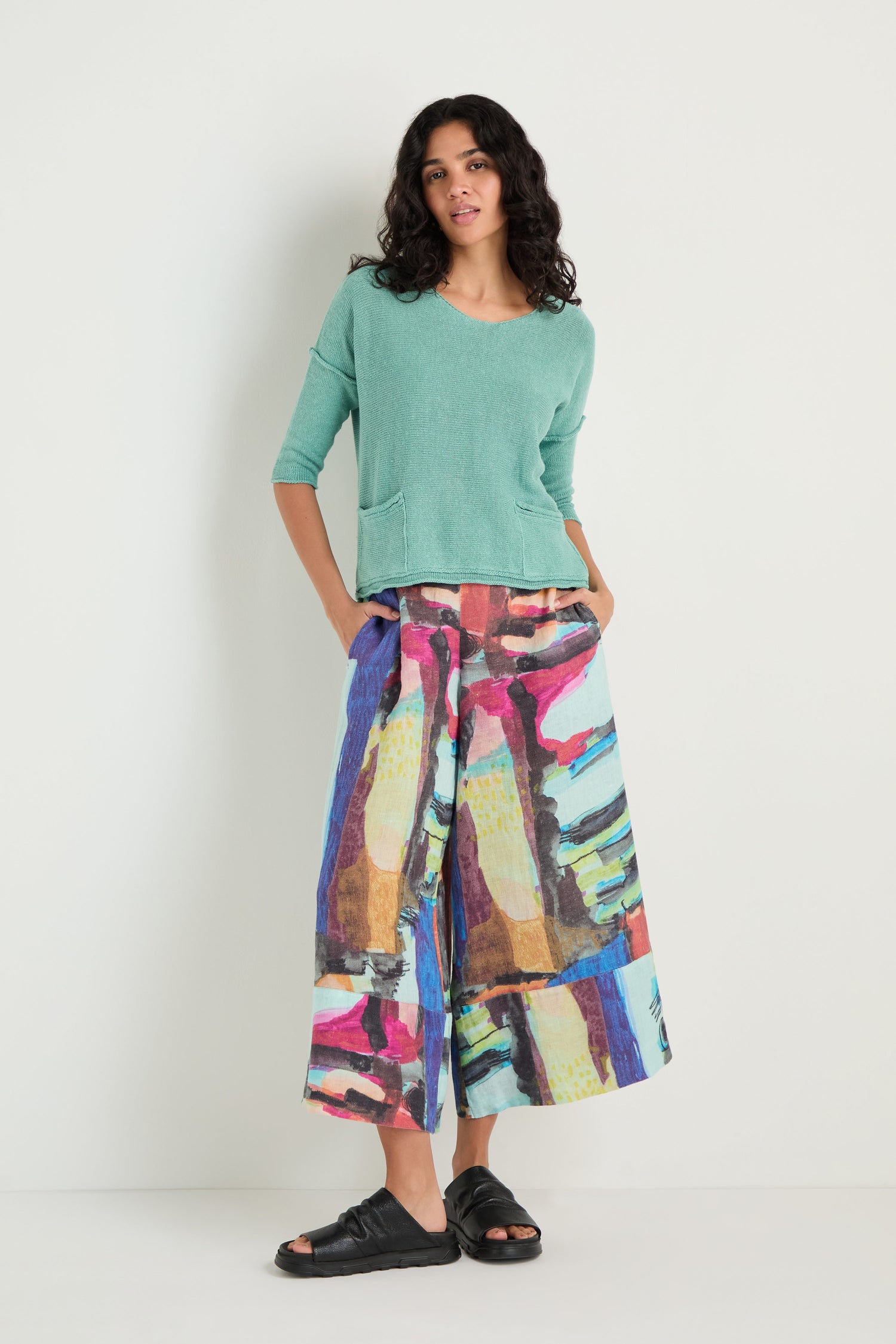 A woman stands against a white wall wearing the Two Pocket Linen Knit Top in teal, colorful abstract wide-leg pants with front pockets, and black sandals.