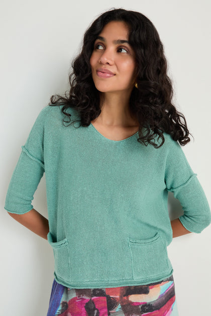 A woman with long curly hair wears the Two Pocket Linen Knit Top in a relaxed silhouette, styled with a colorful skirt, standing against a plain light background.
