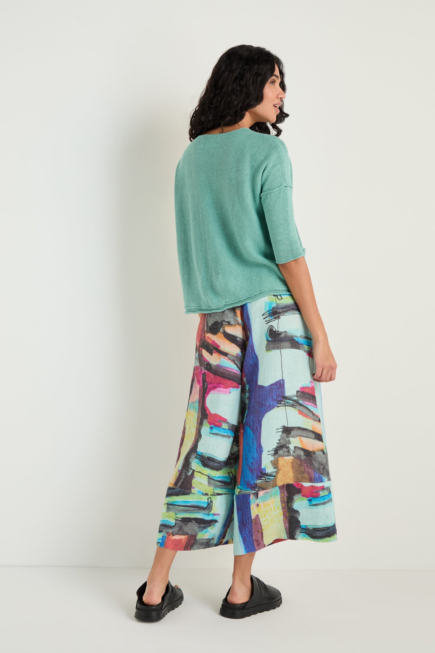 Woman stands against a plain wall, shown from the back, wearing a loose green Two Pocket Linen Knit Top, colorful abstract wide-leg pants with front pockets, and black slip-on shoes.