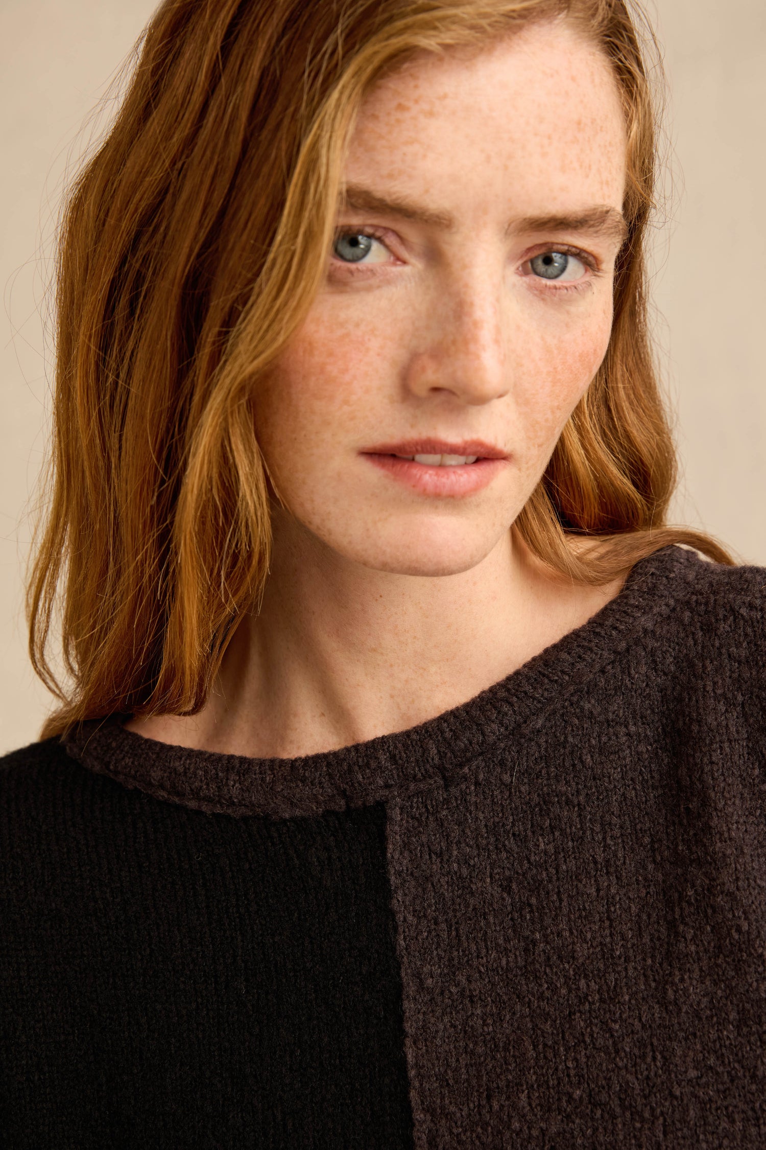 A woman with long red hair and blue eyes wears the Two Colour Knit, a dark, textured sweater with an asymmetric design. She gazes directly at the camera with a neutral expression.