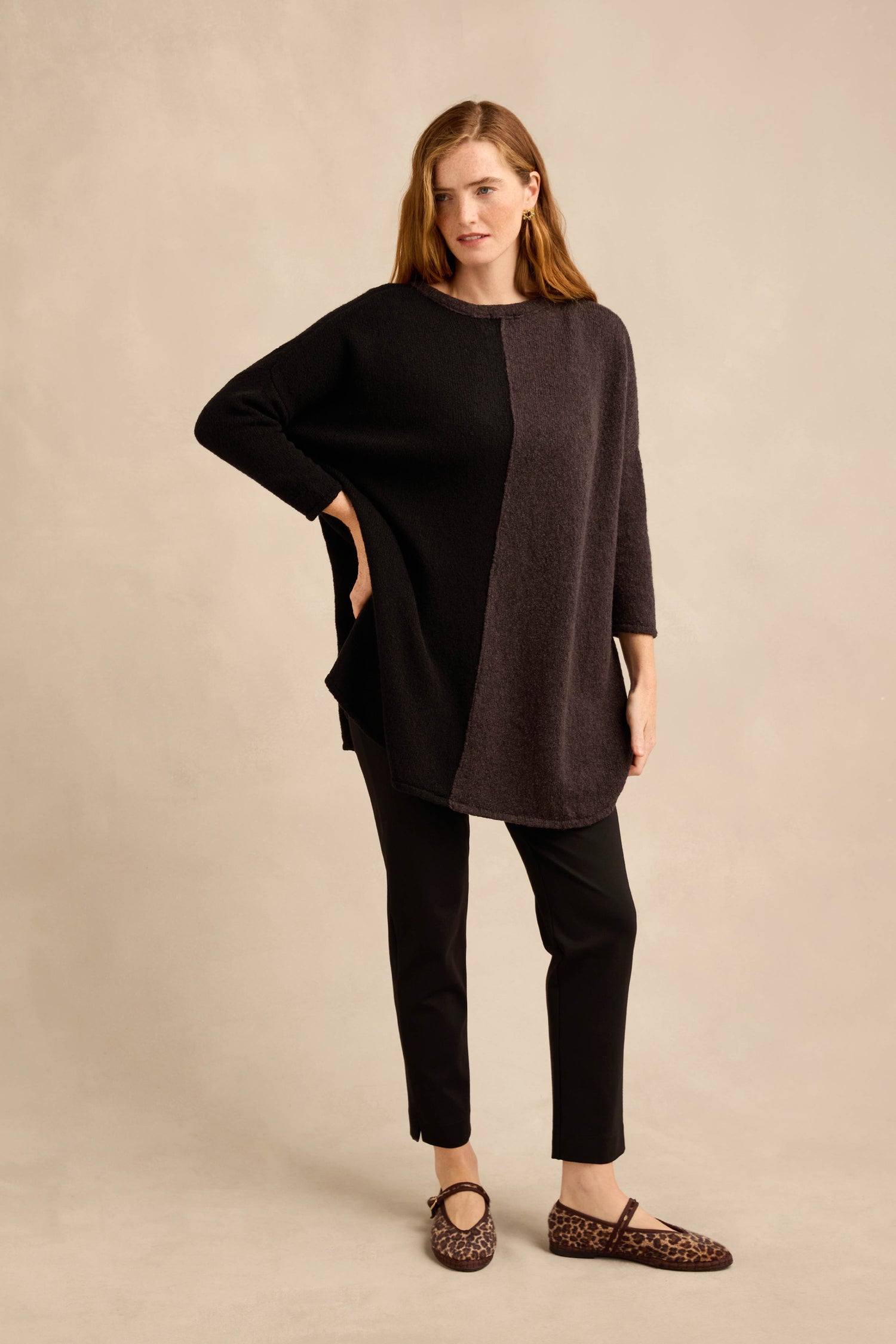 A woman stands posing in the Two Colour Knit, a wool-rich tunic featuring an asymmetric design in two-tone black and dark gray, paired with black pants and leopard print shoes against a beige background.