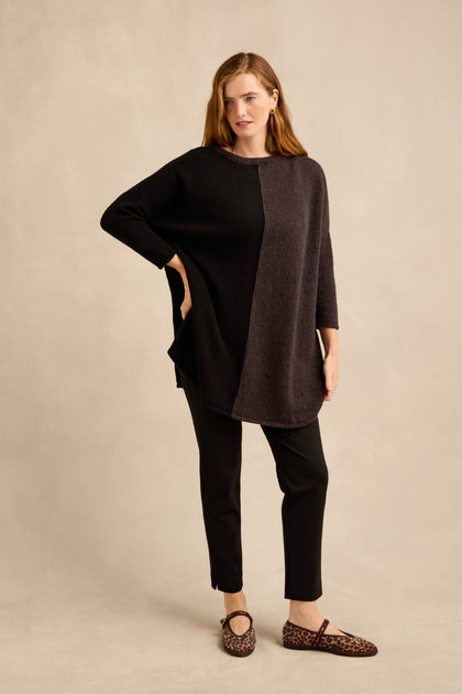 A woman stands posing in the Two Colour Knit, a wool-rich tunic featuring an asymmetric design in two-tone black and dark gray, paired with black pants and leopard print shoes against a beige background.