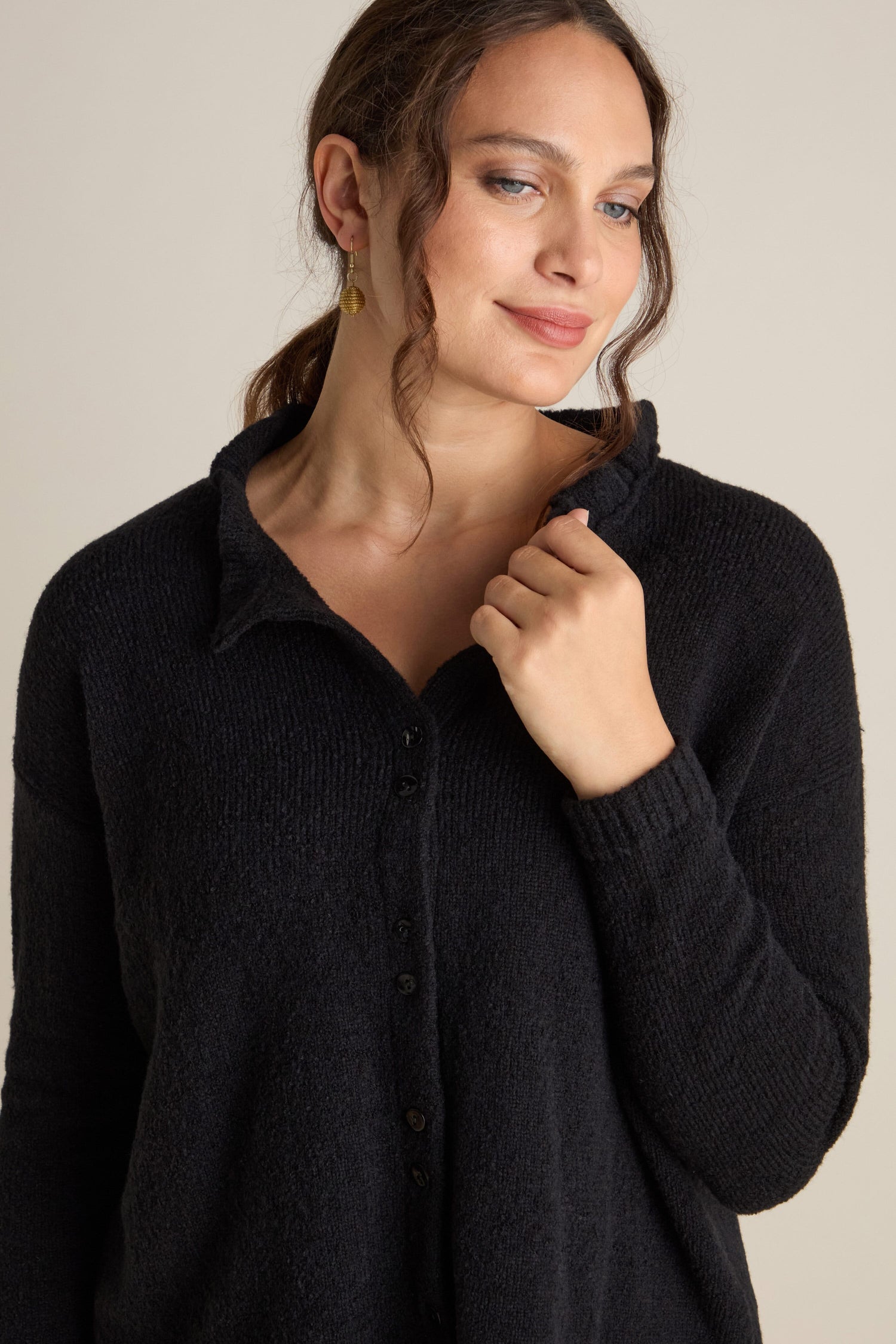 A woman styled in the Frill Neck Wool Cardi and gold hoop earrings is pictured against a plain background. She gently grasps one side of the cardigan’s collar with her right hand and gazes slightly to the side, showcasing a relaxed, effortless look.