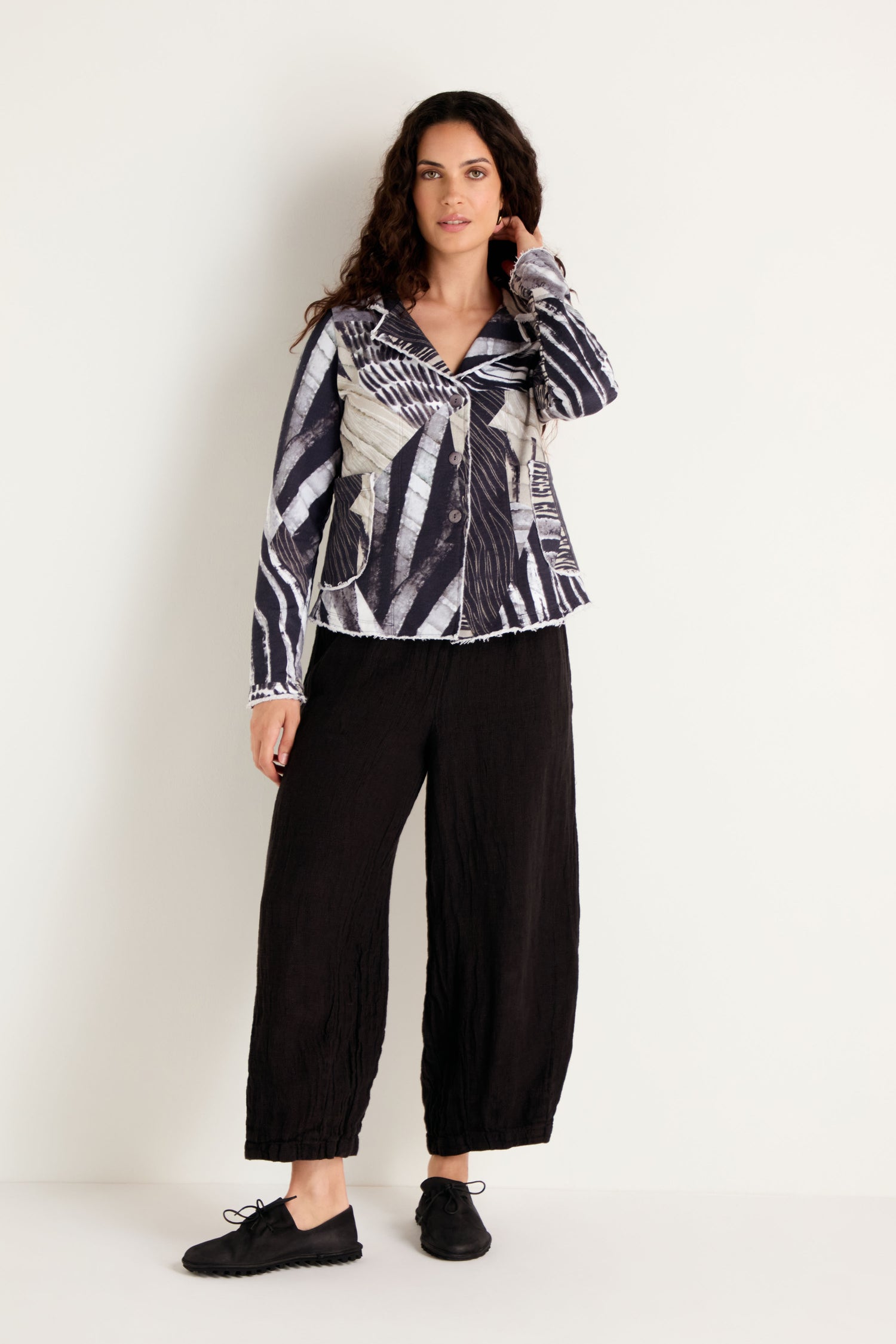 A woman stands against a plain wall, wearing the Monochrome Patchwork Jersey Jacket, paired with black wide-leg pants and black shoes, with one hand touching her hair.