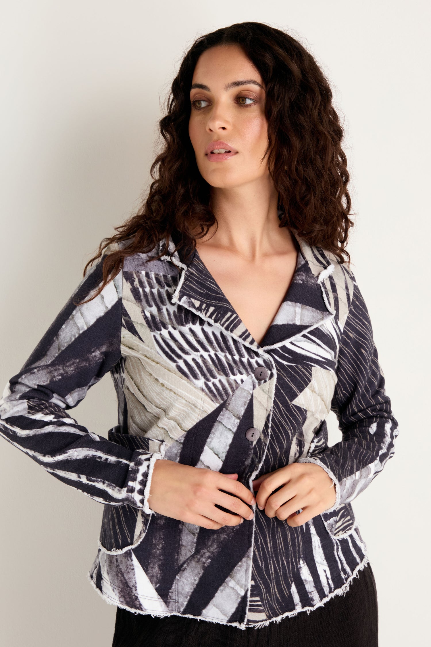 A woman with long curly hair stands against a plain light-colored background, looking to the side as she adjusts her Monochrome Patchwork Jersey Jacket.