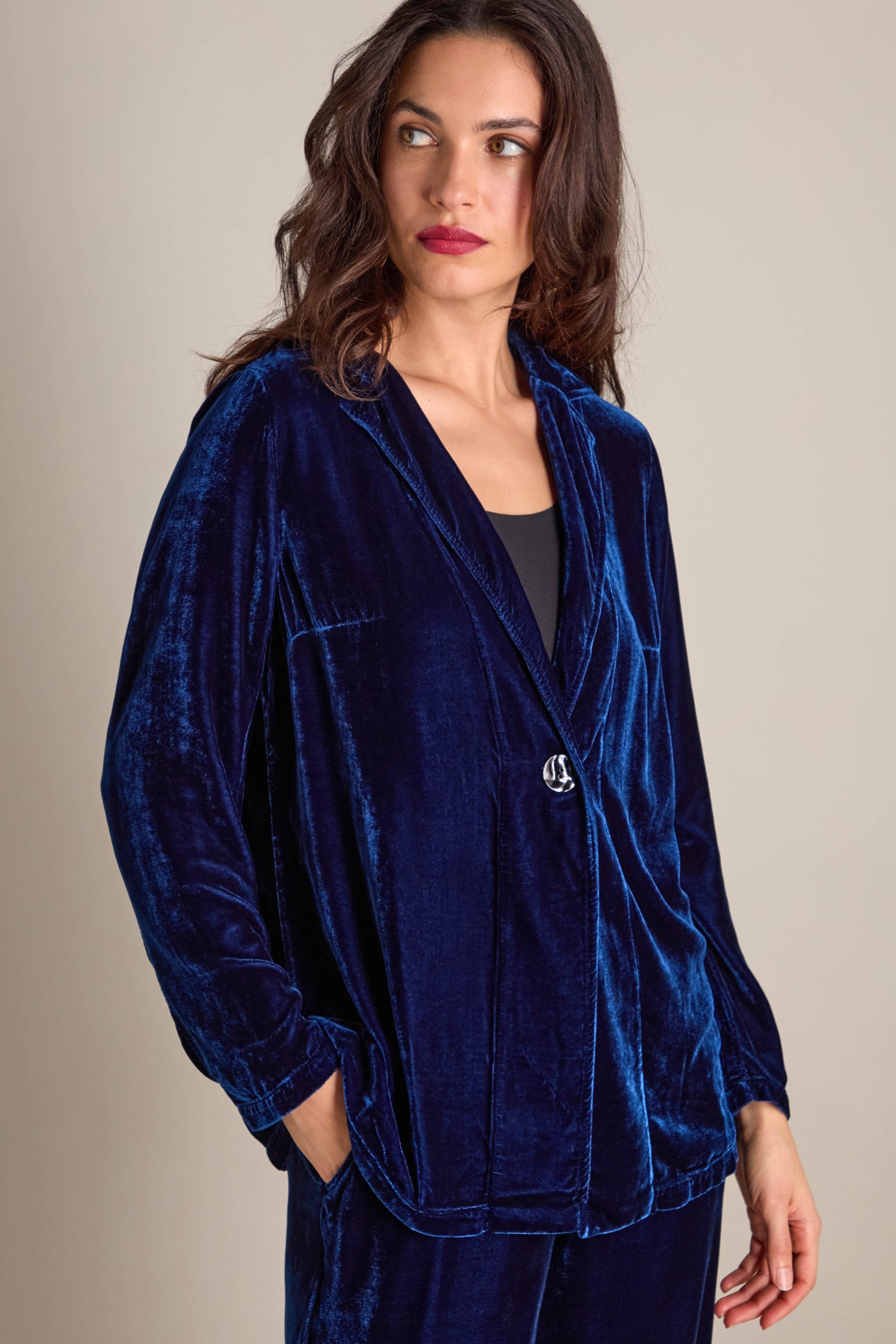 The woman stands elegantly, adorned in the Boxy Silk Velvet Jacket in blue, featuring a single button. The modern sophistication of its boxy silhouette enhances her thoughtful gaze to the side.