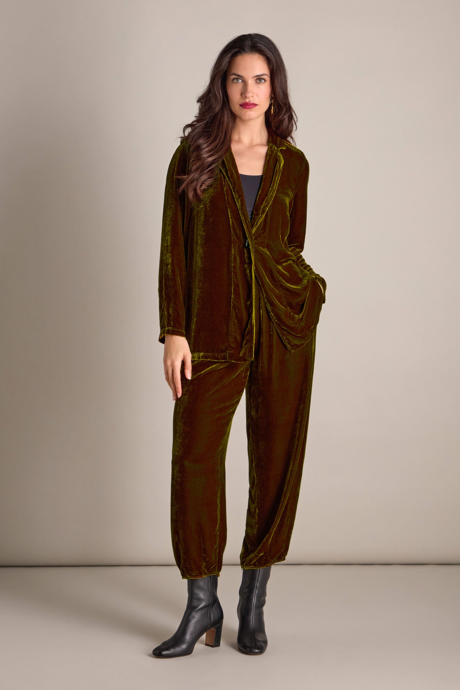 A person in a green Boxy Silk Velvet Jacket and black boots poses gracefully against a neutral background, highlighting their stylish ensemble.