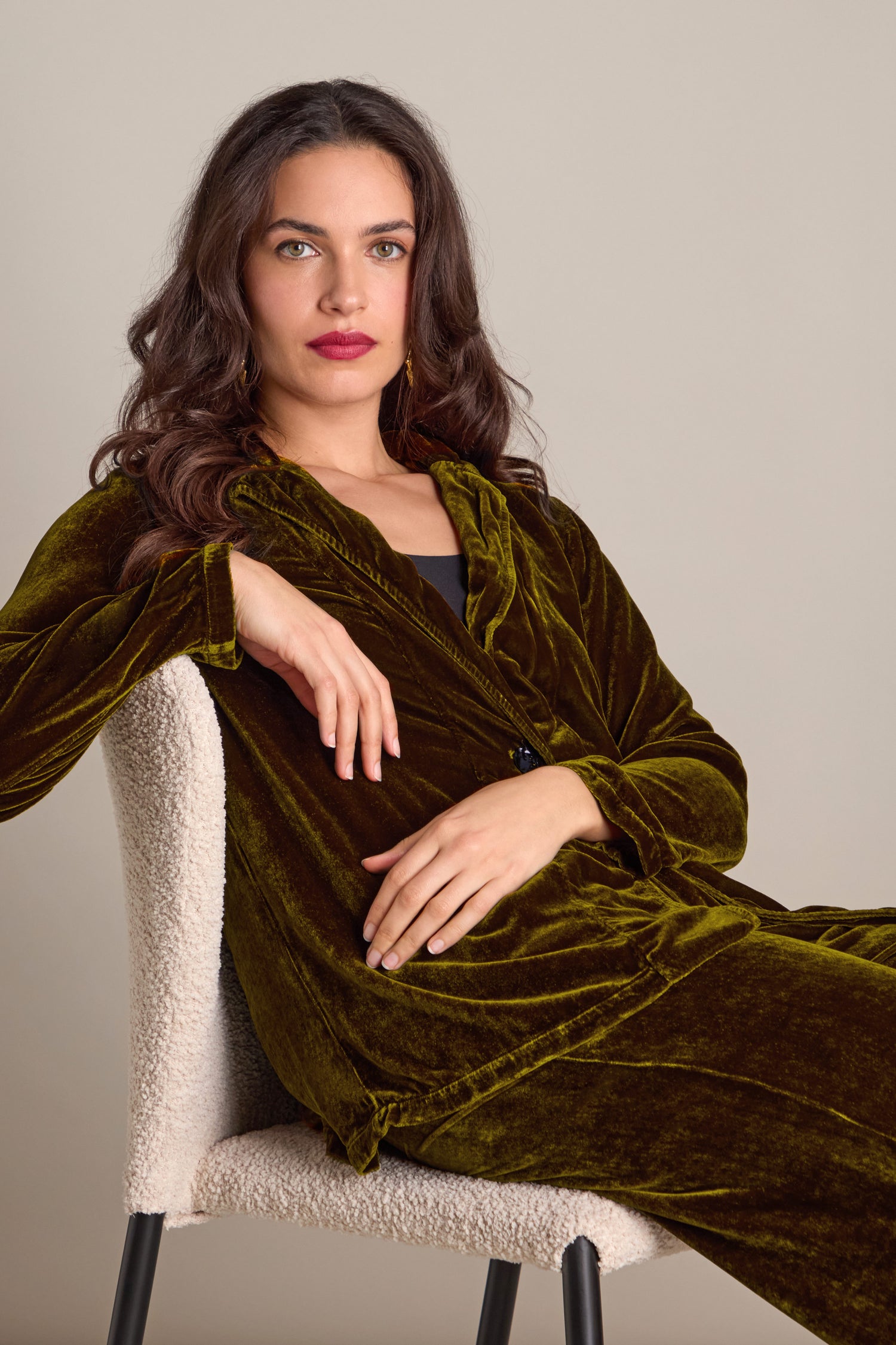 A woman is seated on a chair, wearing the Grizas Boxy Silk Velvet Jacket in dark green, looking at the camera with a neutral expression.