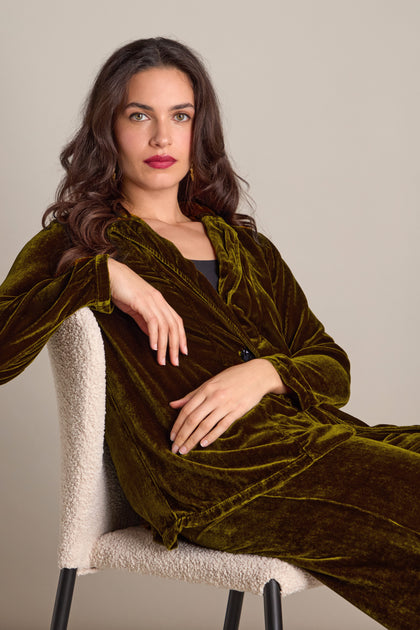 A woman is seated on a chair, wearing the Grizas Boxy Silk Velvet Jacket in dark green, looking at the camera with a neutral expression.