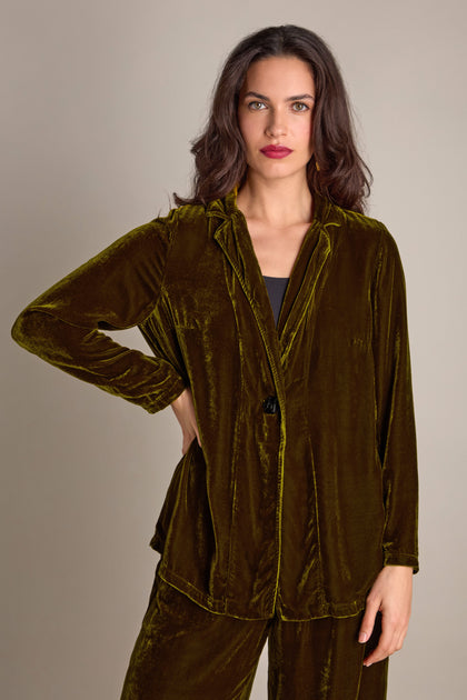 A woman in a boxy silk velvet jacket from Grizas in an olive-green hue stands against a plain background, looking directly at the camera with a neutral expression.