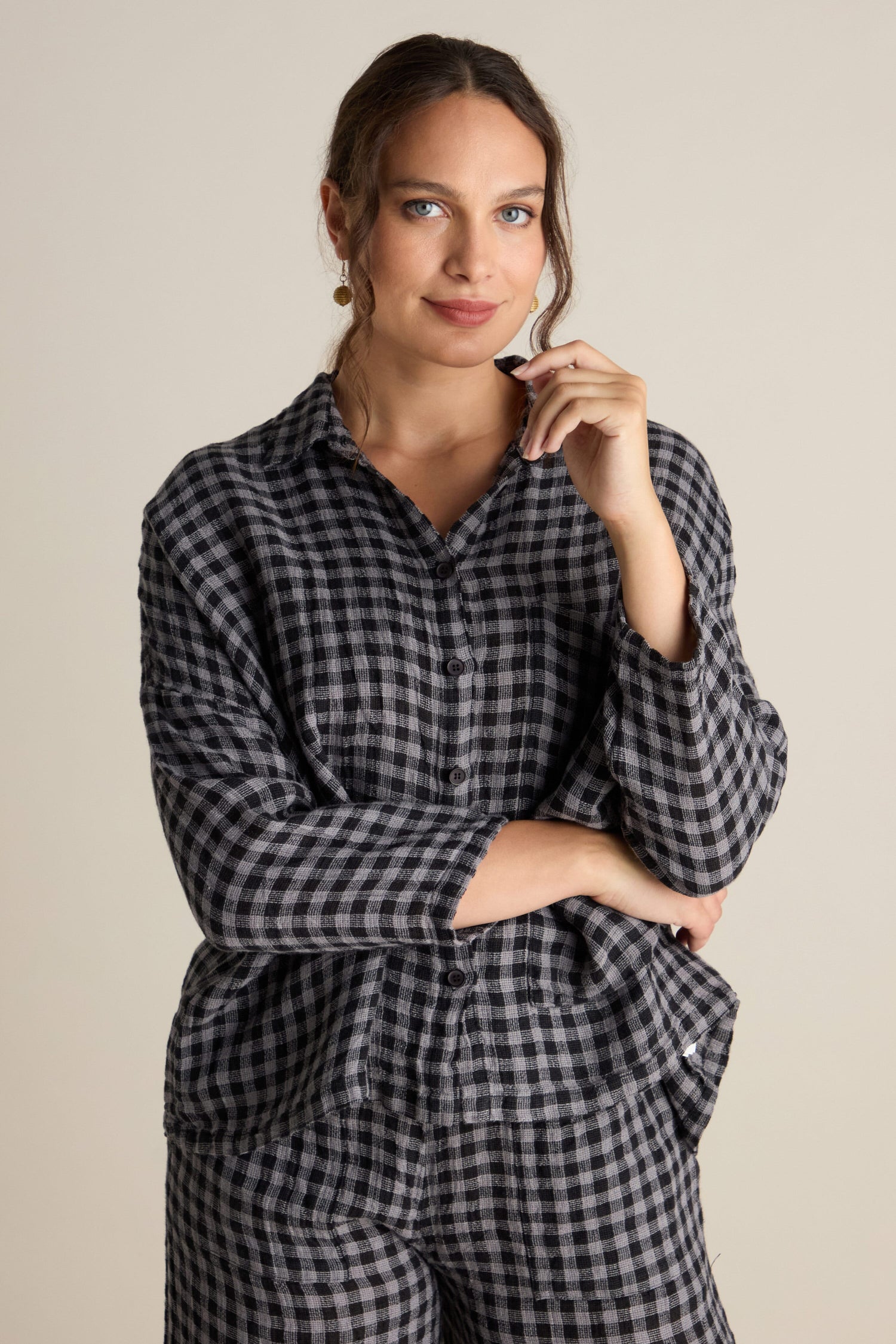 Woman with long hair wearing the Linen Check Big Pocket Shirt and matching pants, standing against a plain background with one hand resting on her chin, showcasing effortless day-dressing.