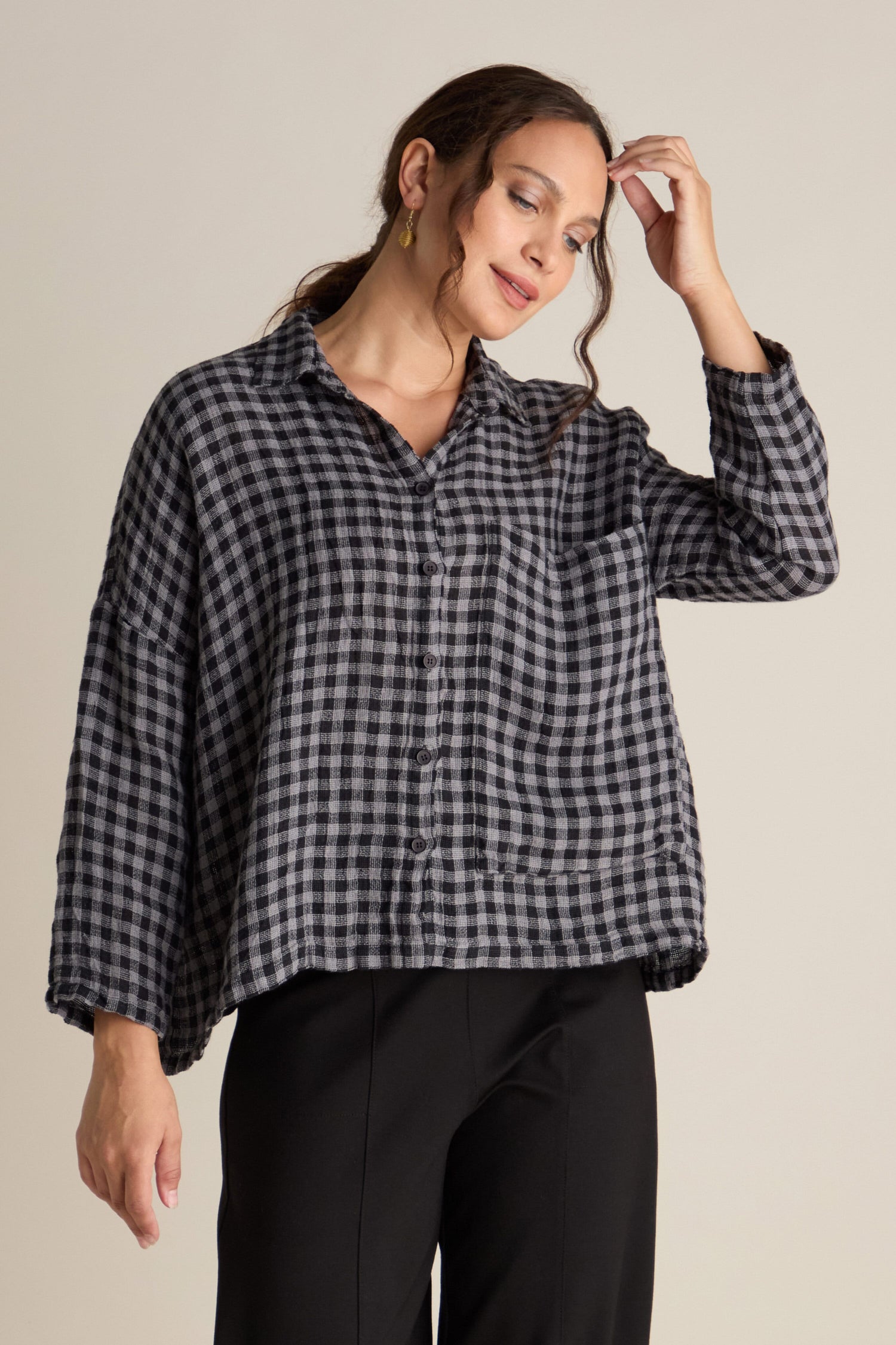 A person wearing the "Linen Check Big Pocket Shirt" in black and gray with an oversized pocket, paired with black pants, stands against a plain background, touching their hair with one hand and wearing earrings.