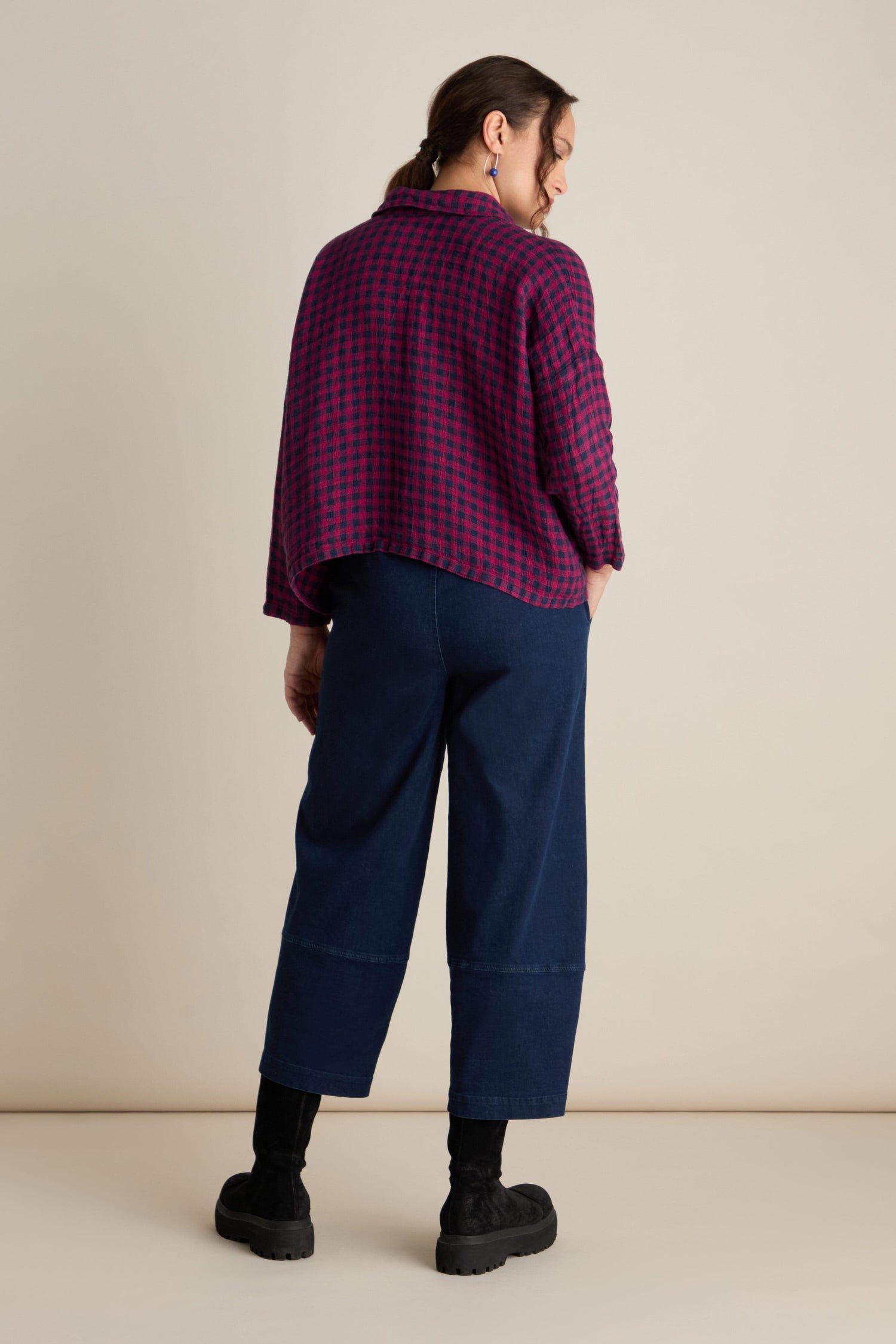 A person stands with their back to the camera, wearing a Linen Check Big Pocket Shirt in red and blue, dark blue wide-leg pants, black socks, and black shoes. Their hair is tied back in a ponytail, perfect for day-dressing.