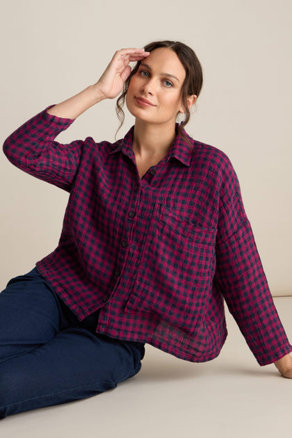 A person with dark brown hair in a low bun, dressed in the Linen Check Big Pocket Shirt featuring a red and black plaid design, and blue pants sits with one hand raised to their face against a plain background.