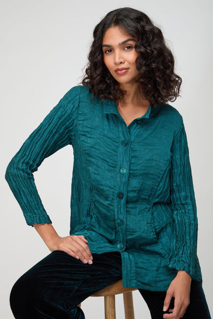 Woman with shoulder-length curly hair wearing a textured teal Panel Front Silk Linen Jacket and black pants, seated on a wooden stool against a plain background.
