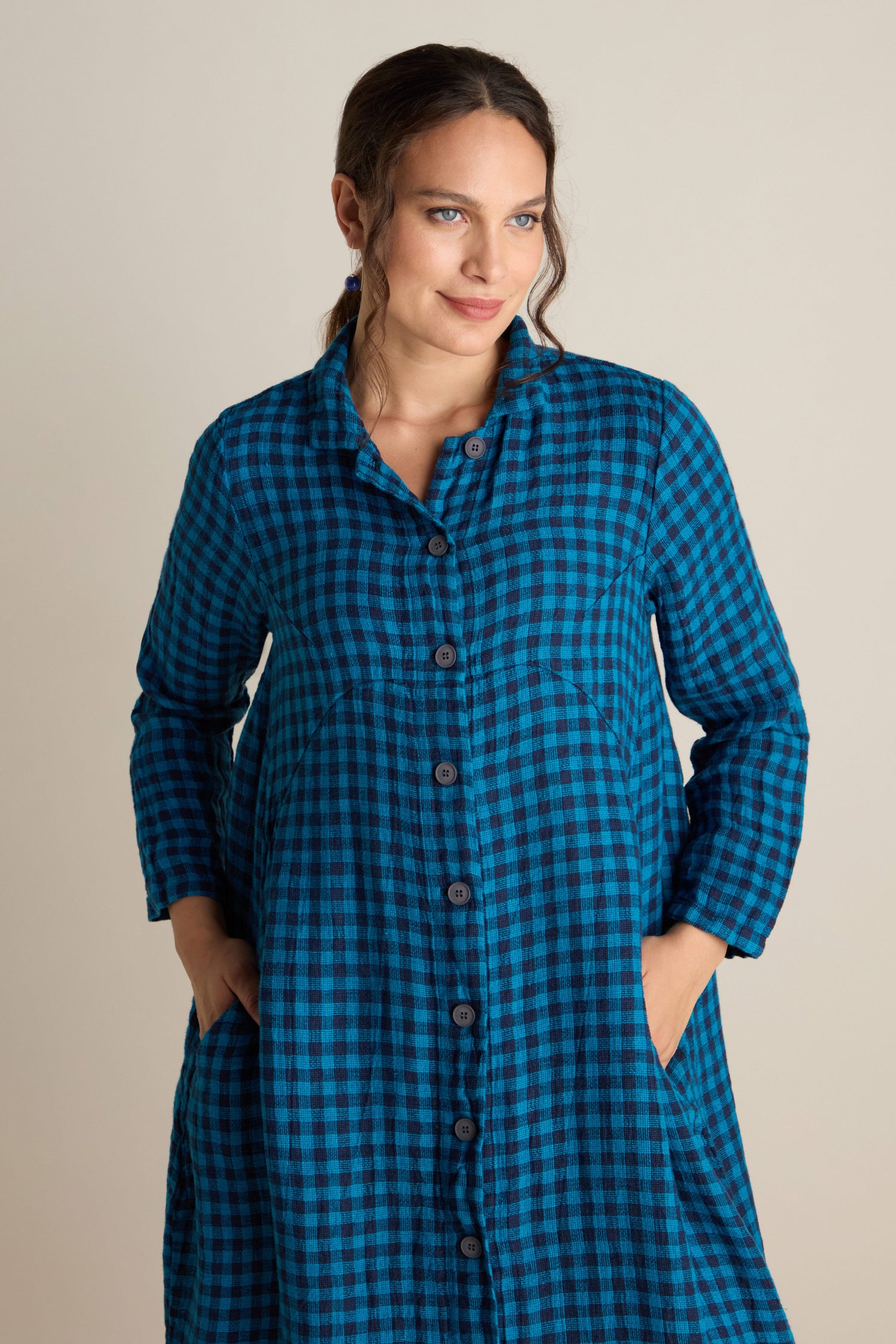 A person is standing against a plain background, wearing a Linen Check Shirt Dress in blue and black checkered pattern, with hands in pockets and a slight smile.