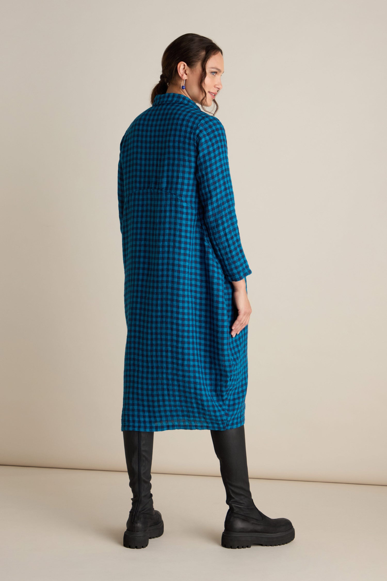 Woman wearing a blue and black checkered long coat, black leggings, and black boots, facing slightly to the side in a neutral studio setting. The look is effortlessly elevated with a loose-fitting Linen Check Shirt Dress layered underneath.