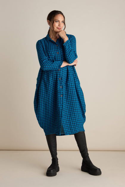 A person stands wearing the Linen Check Shirt Dress, paired with black leggings and black boots. They have one hand resting under their chin and gaze off to the side against a plain, neutral-colored background.